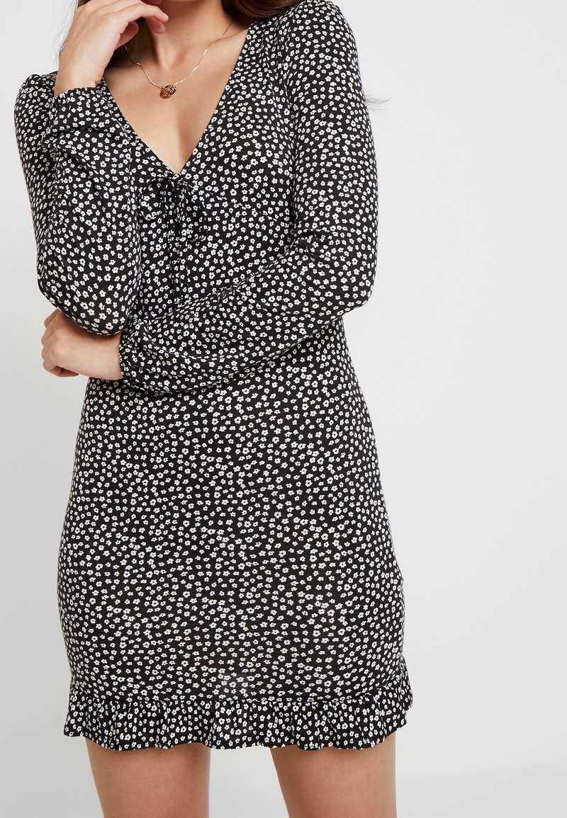 Czarna sukienka z białymi wzorami kwiatowymi, dekolt w serek, długie rękawy i falbaniasty dół. Wykonana z lekkiego materiału o dopasowanym kształcie.