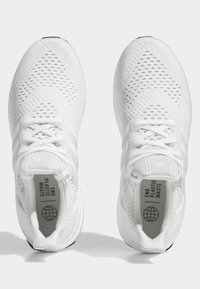 Vita idrottsskor med ovandel i mesh, texturerad design, framträdande snörning och gummisula med "END PLASTIC WASTE" tryckt inuti.