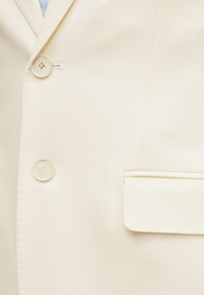 Σακάκι κοστουμιού σε κρεμ απόχρωση με δύο ορατά κουμπιά, μία τσέπη με καπάκι και μέρος ενός ανοιχτό μπλε πουκάμισου από κάτω.