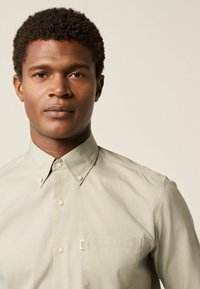 Hellgraues Hemd mit Button-Down-Kragen, Tasche auf der linken Seite und leichtem Stoff. Die Knopfdetails sind sichtbar.