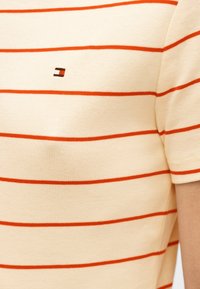 Μπεζ πουκάμισο με κοντά μανίκια, λεπτές οριζόντιες κόκκινες ρίγες και ένα μικρό ορθογώνιο λογότυπο κόκκινου, λευκού και μαύρου χρώματος στο στήθος.