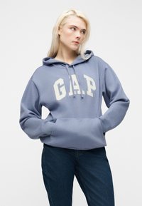 Ξανθιά γυναίκα που φοράει μπλε φούτερ GAP με κουκούλα, με τα χέρια στην μπροστινή τσέπη, κοιτάζοντας προς το πλάι, συνδυασμένο με σκούρο μπλε τζιν, σε λευκό φόντο.