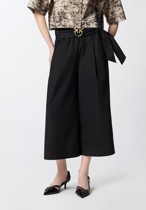 Donna che indossa pantaloni neri a gamba larga e corte con vita con cintura e décolleté nere con punta a punta, abbinati a una maglietta a maniche corte con stampa a motivo di serpente.