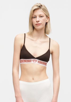 Tânără cu păr blond, purtând un bralette maro închis cu model roșu cheie grecească și logo Medusa pe o bandă albă, asortat cu pantaloni albi.