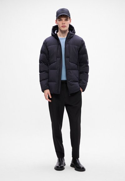 Veste en doudoune noire avec capuchon, comportant de grandes sections matelassées. Portée sur un haut bleu clair et un pantalon noir ajusté avec des bottes noires.