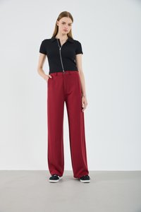 Woman wearing a black short-sleeve zip-up top and wide-leg red pants, standing with one hand in pocket and black sneakers on gray floor.