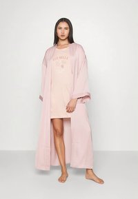 Lang, lys rosa kappe med vide ermer, båret over en kort rosa kjole med bladdesign og tekst. Jevn, myk stoffstruktur.