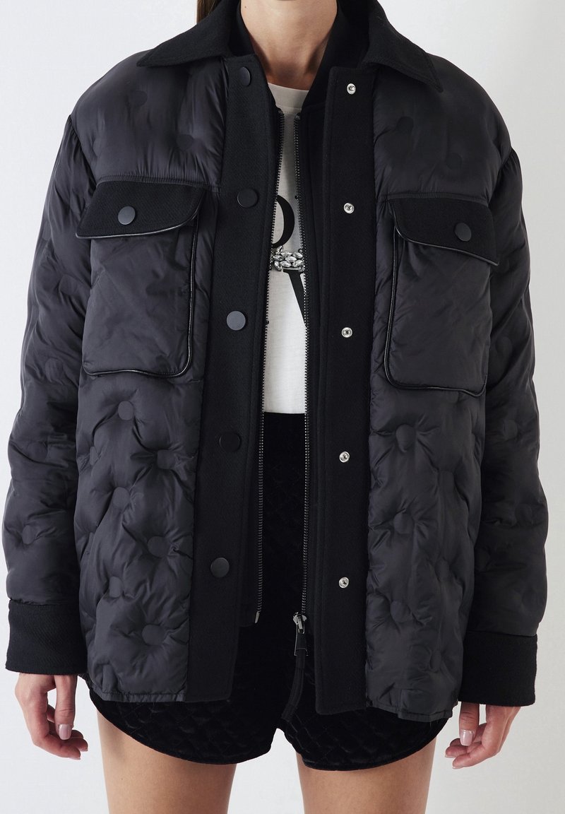 Veste matelassée noire avec grandes poches à rabat et boutons pression, portée sur un haut blanc à motif graphique et un short matelassé noir.