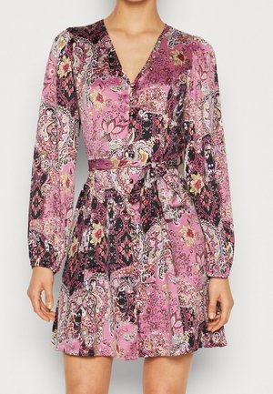 Vrouw draagt een langermige, getailleerde jurk met V-hals en een roze, zwarte en gele bloemen- en paisleyprint.
