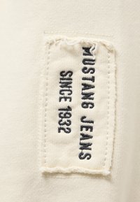 Ausgefranster rechteckiger Stofflabel, auf cremigem Strick genäht, mit schwarzem Text, der "Mustang Jeans Seit 1932" in vertikaler Orientierung liest.