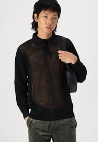 Svart mesh genser med polo-krage og kort placket, parret med mørkegrønne bukser og en liten svart skulderveske.