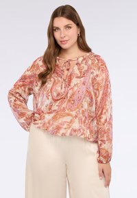 Lehká, vzorovaná blůza v teplých odstínech růžové a oranžové, s dlouhými rukávy, vázacím výstřihem a mírně nabíraným lemem. Kombinována se béžovými kalhotami.