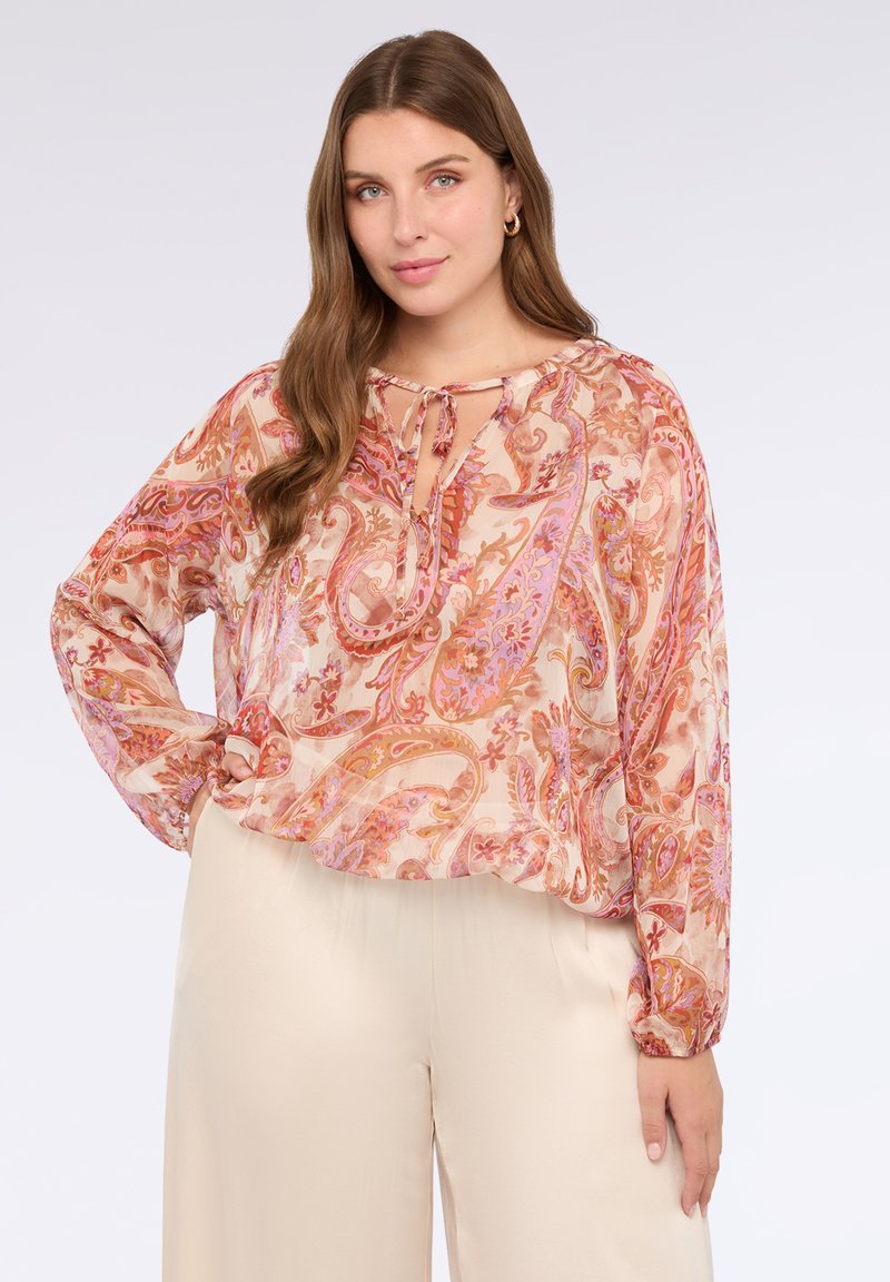 Lehká, vzorovaná blůza v teplých odstínech růžové a oranžové, s dlouhými rukávy, vázacím výstřihem a mírně nabíraným lemem. Kombinována se béžovými kalhotami.