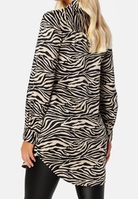 Long-sleeve animal print shirt featuring a beige background with black zebra stripes, a curved hem, and buttoned cuffs. Soft fabric texture.