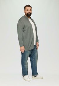 Cardigan gris avec col montant, associé à un t-shirt blanc et un jean bleu. Le mannequin porte des baskets blanches. Design simple et décontracté.