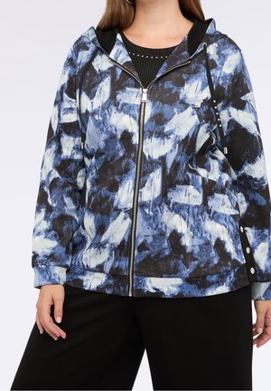 Frau trägt einen Kapuzenpullover mit Reißverschluss mit blau-weiß-schwarzem abstraktem Muster und schwarze Hose, steht mit einer Hand entspannt an der Seite.