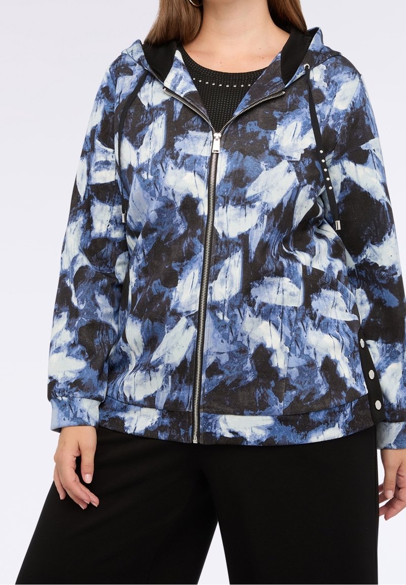 Femme portant un sweat à capuche zippé avec un motif abstrait bleu, blanc et noir et un pantalon noir, debout avec une main détendue sur le côté.