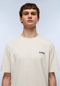 Cremefarvet t-shirt med korte ærmer, rund halsudskæring og et lille sort logo på venstre brystområde. Glat bomuldsstruktur.