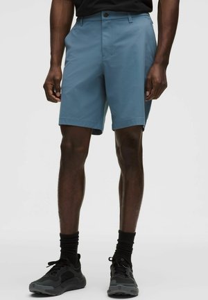 Mand iført blå shorts til midt på låret, sorte sokker, sorte sneakers, stående mod en ensartet lys baggrund.