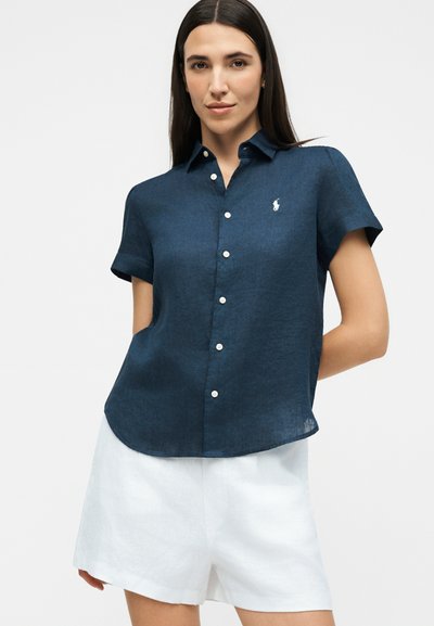 Γυναίκα φοράει μπλε σκούρο κοντομάνικο πουκάμισο με κουμπιά, λευκό κεντημένο λογότυπο και λευκά σορτς με ψηλή μέση, σε απλό φόντο.