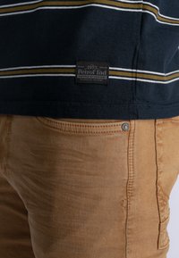 Mörkblå T-shirt med vita och senapsgula ränder, med en svart etikett. Matchas med ljusbruna byxor, som visar textilstruktur och metallaccenter.