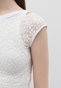 Top bianco in pizzo con un motivo a trama, scollatura rotonda classica e maniche corte. Il tessuto sembra leggero e delicato.