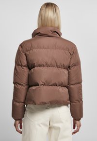 Brun pufferjakke med høj krave, quiltet tekstur og elastiske manchetter. Båret med off-white bukser, der viser et cropped design.