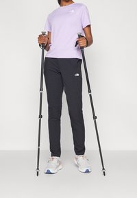 Světle fialová košile s krátkým rukávem, černé sportovní kalhoty a bílé tenisky s oranžovými detaily. Uživatel drží trekové hole, stojí na jednobarevném pozadí.