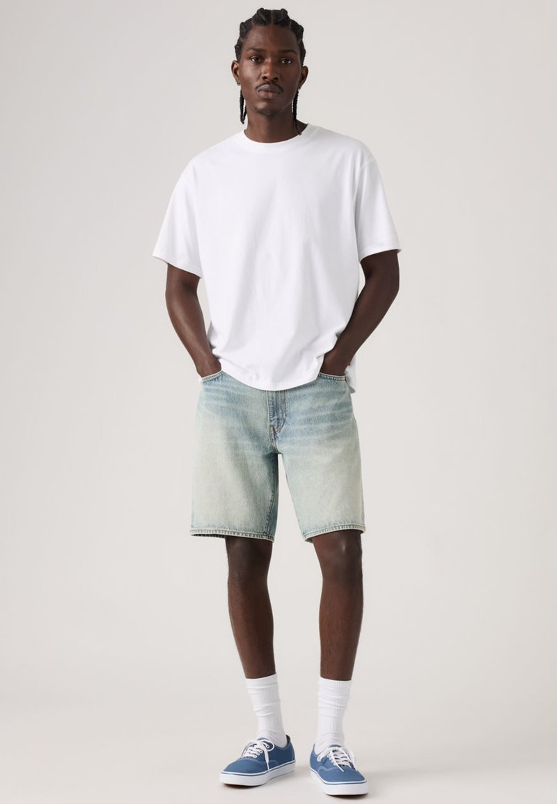 Maglietta bianca di cotone, pantaloncini di denim azzurro con orlo sfrangiato, scarpe da ginnastica blu, calzini bianchi. La modella ha i capelli intrecciati e le mani nelle tasche.