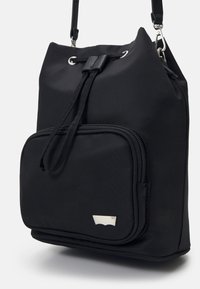 Sort nylon taske med snøre i toppen og en frontlomme med lynlås. Har et sølvfarvet logo og en justerbar skulderrem.