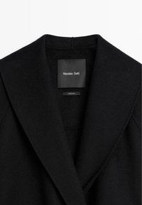 Czarny kołnierz wełnianego płaszcza z czarną metką z logo marki "Massimo Dutti" oraz małą etykietą z napisem "Wyprodukowano w Chinach."