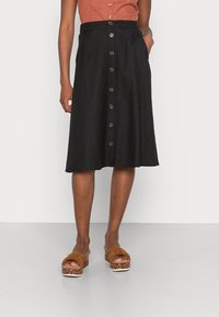 Jupe en denim noir avec une taille ajustée, ourlet au genou, six boutons saillants à l'avant et poches latérales. Portée avec des sandales brunes à nœud.