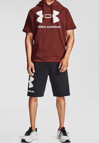 Czerwona bluza z kapturem z dużym białym logo, w połączeniu z czarnymi szortami z białym logo. Obuwie to białe sneakersy z czarnymi akcentami.