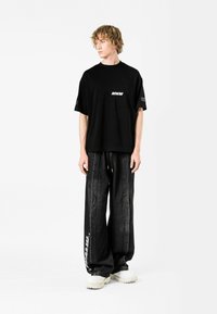 Camiseta negra de manga corta con un bolsillo y el texto "MWM", combinada con unos pantalones vaqueros oscuros de corte holgado que presentan un gráfico lateral y zapatillas blancas.