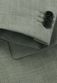 Tissu de costume sur mesure vert avec une texture fine, arborant trois boutons noirs et des détails de revers soigneusement cousus.