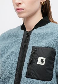 Világoskék polárdzseki fekete részletekkel, cipzáras elülső résszel, fekete folt zsebbel és textúrázott anyaggal. Közeli nézet.