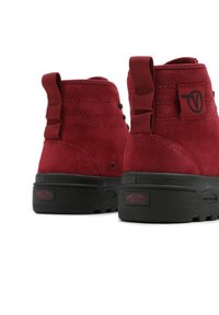 Bottes en daim rouge avec un design montante, dotées d'une semelle en caoutchouc noire et d'accents texturés, incluant un patch logo et des détails de couture.