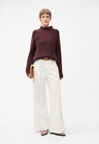 Maglione a collo alto bordeaux lavorato a maglia, pantaloni bianchi larghi, cintura marrone, tiene una pochette color sabbia. Le scarpe sono grigie chiare con punta a punta.