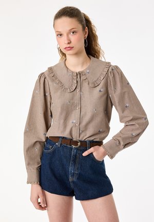 Jonge vrouw draagt een beige gestreepte blouse met een grote gerimpelde kraag en spijkershort met een bruine riem, staand met één hand in de zak.