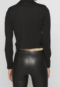 Zwarte cropped jas met lange mouwen, gemaakt van een soepele stof. Kenmerkt zich door een achterriem en minimalistisch ontwerp, gecombineerd met hoogglanzende zwarte broek.