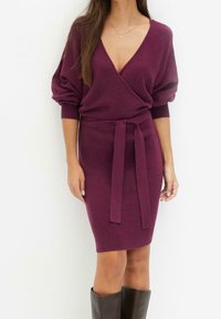Robe en tricot bordeaux avec un décolleté en V profond, des manches bouffantes et une taille nouée. Présente une coupe moulante et une texture côtelée.