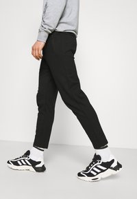 Svarta avsmalnande byxor i slät tyg med sidodragkedjedetalj, i kombination med svarta och vita sportskor samt vita ankelsockor.