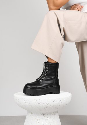 Botines de cuero negro hasta el tobillo con una gruesa plataforma y un acabado texturizado, que presentan ojetes de metal y una cremallera lateral, exhibidos sobre un pedestal blanco.