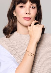 Złota bransoletka z zawieszkami w kształcie gwiazdy i serca, złożona z ogniw. Noszona na nadgarstku w zestawieniu z teksturowanym beżowym swetrem.