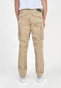 Man in beige cargobroek en witte sneakers, staand met de rug naar de camera tegen een plain witte achtergrond.