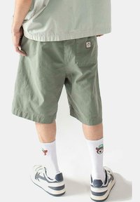 Shorts de algodón verde con un corte relajado, a la altura de la rodilla, que cuentan con un bolsillo trasero con un parche de logo, combinados con calcetines blancos y zapatillas deportivas.