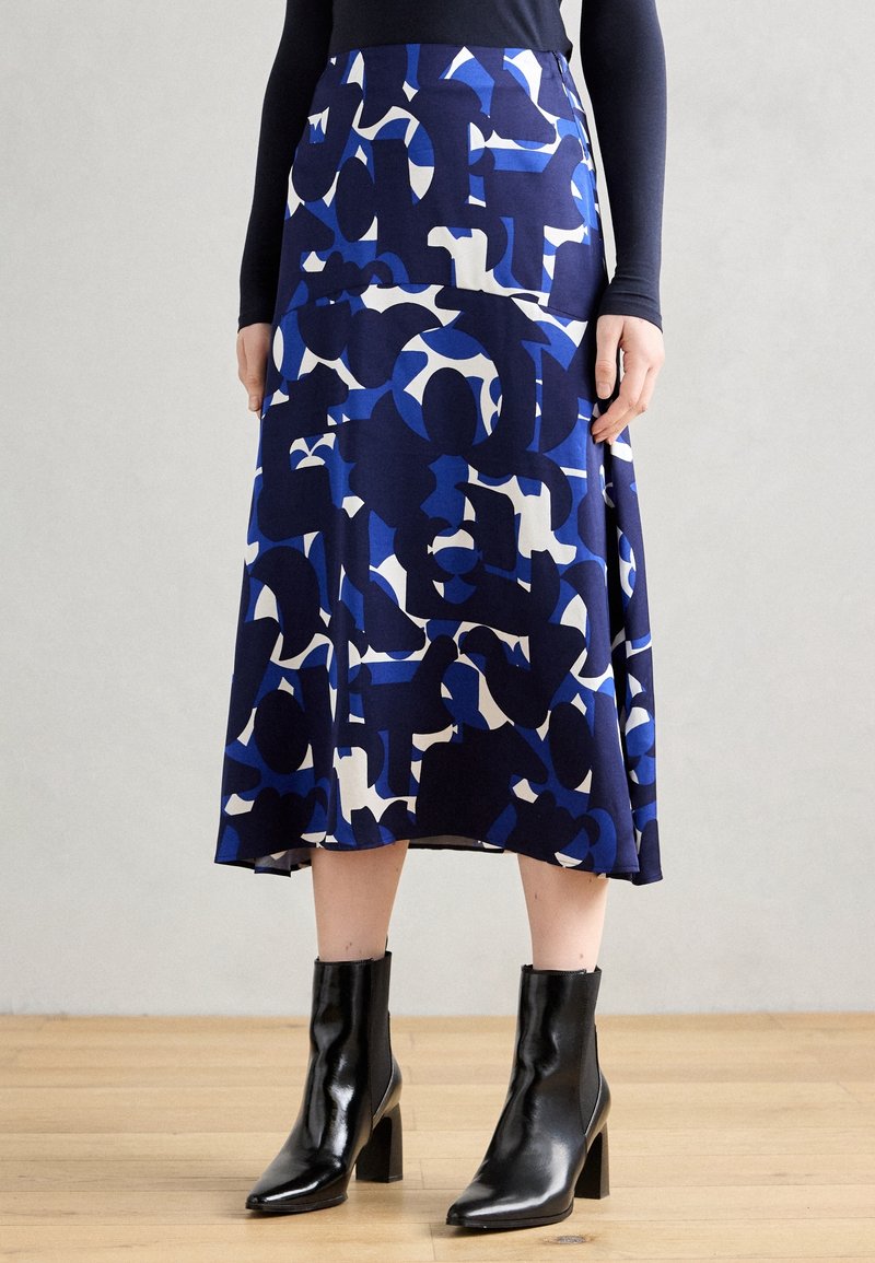 Femme portant une jupe midi à motif abstrait bleu et blanc, un haut noir à manches longues, et des bottines noires à talons, debout sur un sol en bois.