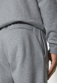 Graue Sweatpants aus weichem Material, mit einer Gesäßtasche und einem dezenten Logo-Detail in Pink. Elastischer Bund für ein angenehmes Tragegefühl.