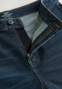 Mörkblå denimjeans med en öppen dragkedja, synlig knapp och etikett "Next Performance Relaxed Fit" inuti midjebandet.