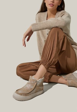 Scarpe beige slip-on con una brillantezza metallica, fodera morbida e suola in gomma texturizzata. Abbinato a pantaloni marroni con fondo arricciato e un maglione leggero.